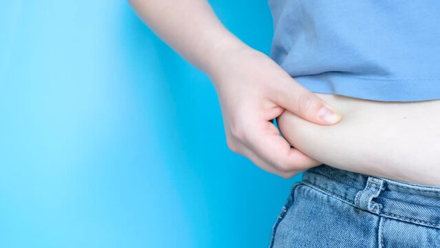 Female hand holding excessive belly fat on blue background, overweight concept