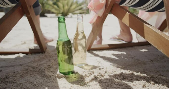 Happy senior caucasian couple sitting in deckchairs on beach reaching for beers, in slow motion