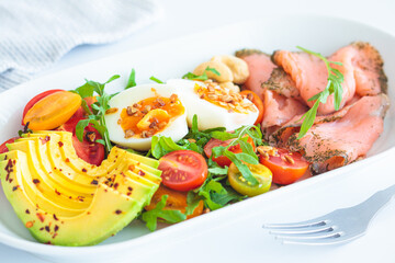 Keto breakfast of salmon, eggs, avocado and nuts on white plate, white background.