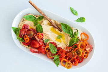 Hummus with tomatoes, peppers, basil and olive oil in white plate, white background.