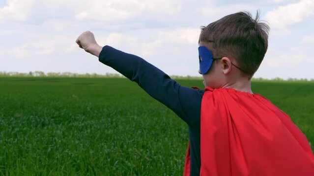 A superhero boy in a red cape and mask raises his hand to the blue sky with clouds. The kid looks at the sky in profile against the background of a green meadow.