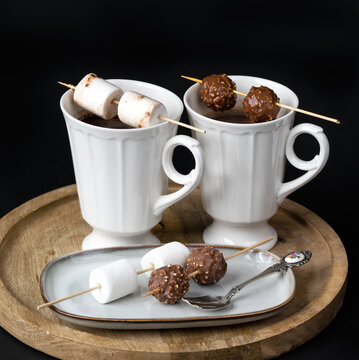 Two beautiful white porcelain cups of hot Chocolate drink with marshmallows and chocolates skewers over wooden table on black background. Winter time. Holiday concept..