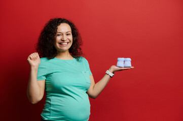 Excited ethnic pregnant woman with cute knitted blue baby booties for her newborn baby, expressing...