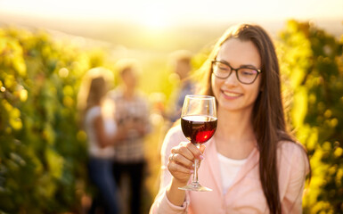 Portrait of a young, millennial woman holding a glass of organic bio red wine outdoors in a vineyard with her friends in the background - Vine-growing, and wine-tasting concept