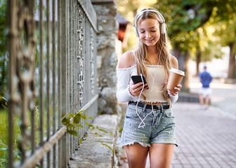 Cheerful teenager student girl walking on a street with a coffee to go paper cup in her hand and listening to music with headphones from a smart phone
