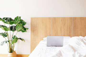 Laptop lying on bed with white linens in bedroom. Modern hotel or home interior with potted monstera plant. Mockup with copy space.