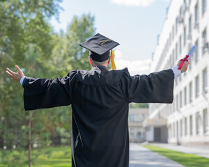 Elderly male graduate rejoices at graduation outdoors. Rear view of a man in a robe with arms...