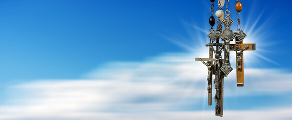 Close-up of a group of metal and wooden crucifixes and rosary beads, against a blurred clear blue sky with clouds and copy space. Photography.