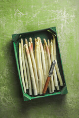 Fresh white asparagus from garden in wooden box with peeler at green rustic background, top view