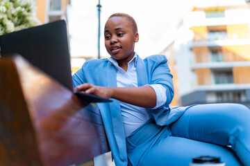 Beautiful young girl in business attire sitting on a bench in front of the office, using a laptop to catch up on unanswered emails