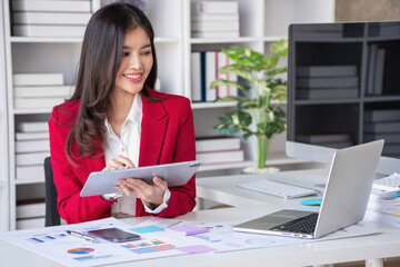 Asian businesswoman in red suit calculating company sales with calculator, laptop, and tablet on table Interior of businesswoman office at the modern workstation.