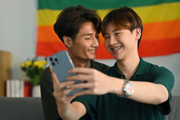 Romantic young gay couple taking a selfie with smartphone in living room rainbow flag in background. LGBT and love concept
