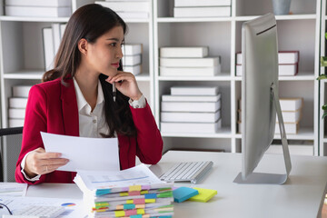 Asian businesswoman in red suit calculating company sales with calculator, laptop, and tablet on table Interior of businesswoman office at the modern workstation.