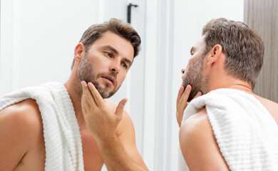 Fototapeta premium Middle aged hispanic man looking in mirror, facial skin and stubble. Male beauty care product. Skincare, home spa. Beauty portrait of a beautiful man. Spa model, moisturizing nourishing creme.