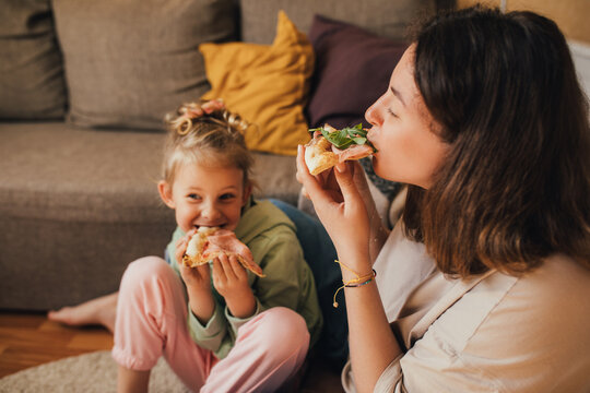 Young mother and her little 5 year old daughter eating pizza together in cozy living room.