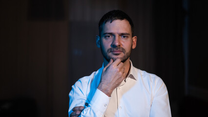 A bearded man stands in the dark and looks ahead puzzled. 