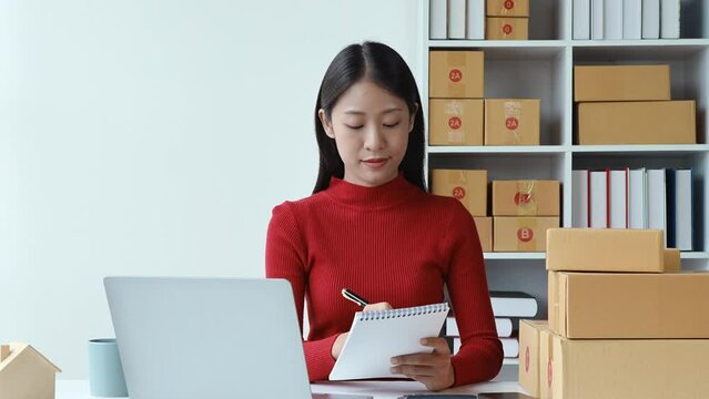 e-commerce, small business, sme , beautiful Asian woman looking at computer and use note taking customer orders and earning from online sales with smiling face happy while working