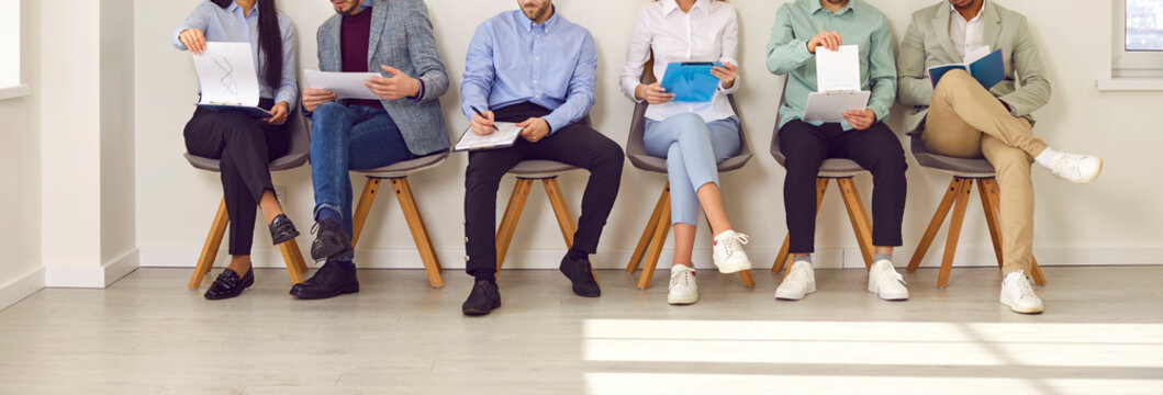 Legs of unrecognizable business people sitting on the chairs in a row with resumes or documents in their hands. Group of a staff. Job candidates seekers waiting for interview invitation turn. Banner.