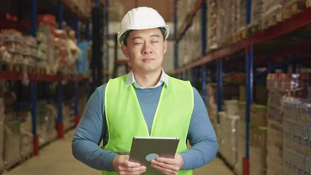 Portrait of handsome asian man wearing safety helmet and reflective vest using digital tablet for work at factory warehouse. Male supervisor inspecting inventory with help of modern gadget.