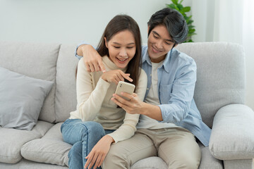 Asian couple smiling and happily use smartphone to online shopping. Husband ready to pay birthday present for beautiful wife. Find accommodation online for honeymoon, App online, book flight tickets.