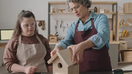Young girl with Down syndrome in apron and protective eyewear polishing wooden plank with sandpaper while making birdhouse with female teacher during woodworking workshop