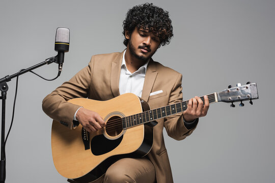 Young and curly indian host of event playing acoustic guitar near microphone on stand while sitting on chair during performance isolated on grey