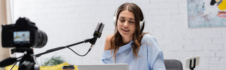 Positive brunette broadcaster in headphones talking during stream near microphone, gadgets and...