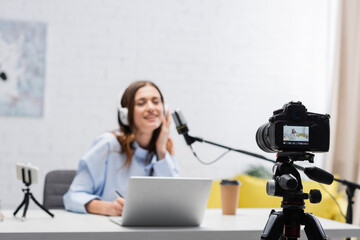 Digital camera near blurred brunette broadcaster in headphones talking during stream near laptop...