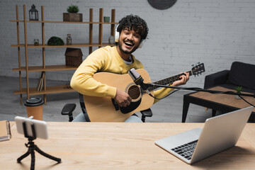 Fototapeta na wymiar Carefree young indian podcaster in wireless headphones playing acoustic guitar near devices and coffee to go in paper cup and notebook on table in podcast studio