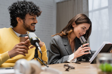 young and curly indian man in yellow jumper smiling near charming colleague looking at notebook and...
