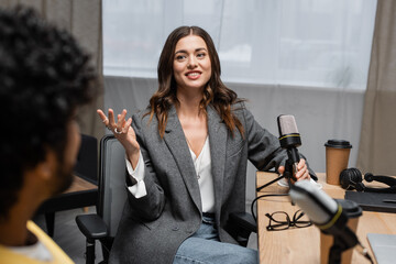 pleased brunette podcaster in grey blazer holding microphone near headphones, notebook, paper cup...