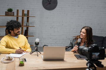 smiling brunette announcer with microphone talking to young and curly indian colleague in yellow...