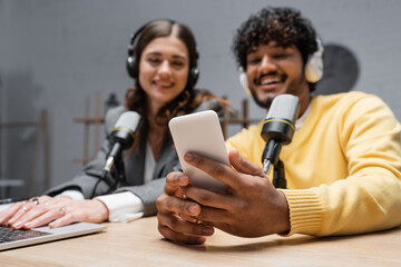 cheerful indian radio host in headphones and yellow blazer holding smartphone near brunette...