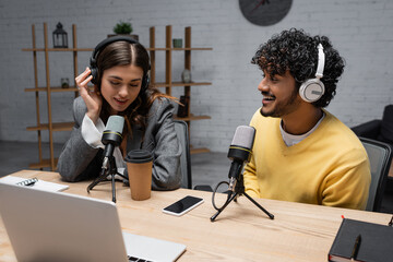brunette podcaster in headphones talking in microphone close to young and happy indian colleague in...