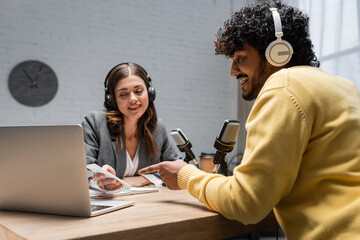 charming radio host in grey blazer showing smartphone to young indian man in headphones and yellow...