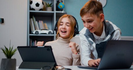 Caucasian boy and girl playing game on laptop and tablet device together with smile face at home in games room. Jojful children sitting the table play online video games and browsing social media in © serg