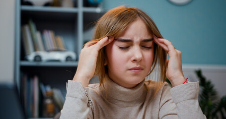 Close up face of tired preteen girl sitting by table with headache massaging her temples with circular motion after a hard day on background bookcase at home