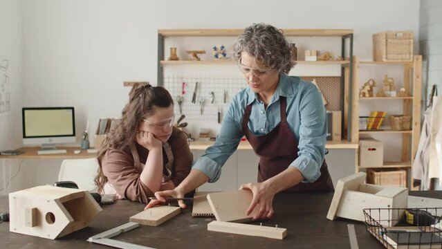 Mid-aged female woodworker in apron and eyeglasses drawing on paper and talking to girl with Down syndrome while teaching her during masterclass in carpentry workshop