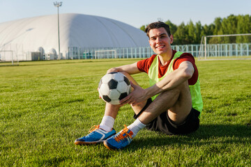 Portrait of a smiling young man football player sitting on field with soccer ball