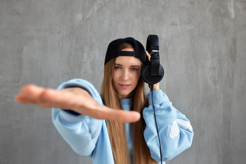 A young pretty long-haired DJ girl in a blue sweater and a black baseball cap holds headphones in her hand and imitates the movements of a hip-hop musician. Music, fun and beauty.