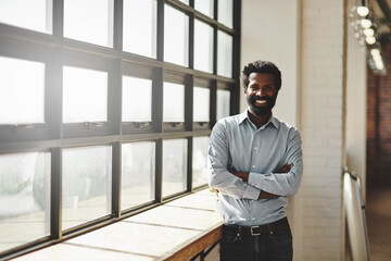 Portrait, window and a business man arms crossed in the office with a smile or mindset of future success. Flare, vision and corporate with a happy male employee standing at work during his break