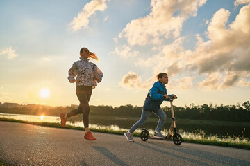 Happy mother and son go in sports outdoors. Boy rides scooter, mom runs on sunny day. Silhouette family at sunset. Fresh air. Health care, authenticity, sense of balance and calmness, mothers Day
