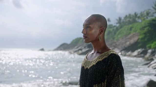 Non-binary black person in boutique dress, golden earrings, nose ring walks on scenic sea beach closeup slow motion. Queer lgbtq pretty fashion model wears jewelry in posh gown in tropical location.