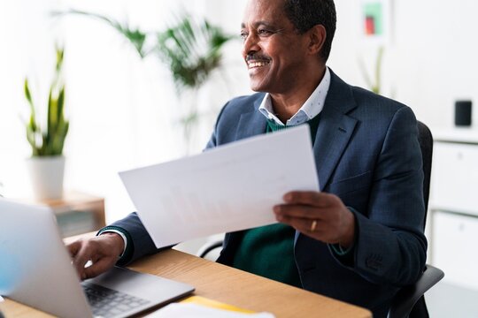 Cheerful elderly Ethiopian male executive manager with documents looking away and smiling while using laptop and working on corporate strategy in modern workspace