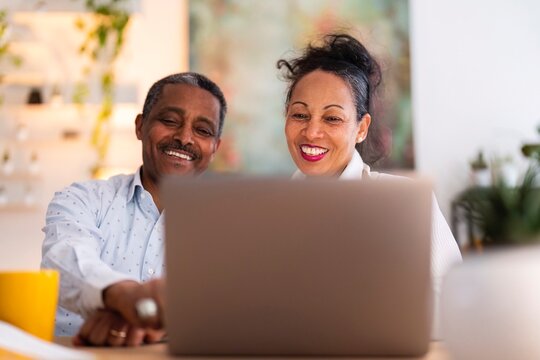 Happy adult Ethiopian couple smiling and doing a video call on laptop together while spending time in light modern apartment