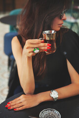 Beautiful glamour woman enjoys glass of Turkish tea alone in cozy cafe, turned and looks away. Stunning fashion model with long dark hair