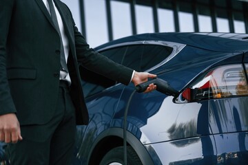 Close up view, putting charger into the socket. Businessman is standing near his electric car outdoors