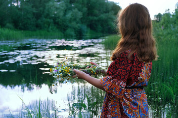 girl with flower wreath in hands near river, abstract natural green background. Esoteric ritual for...