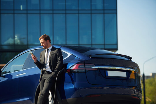 Business building behind. Man in suit is standing near his electric car outdoors