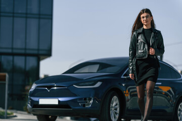 In black jacket and skirt. Young woman is near her electric car outdoors